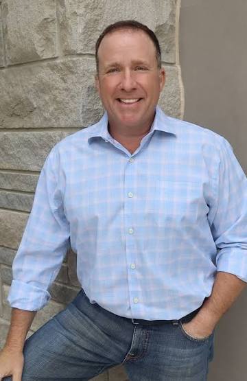 Scott Mandell, advisor, wearing a light blue and white checkered button-down shirt and blue jeans, standing in front of a stone wall.