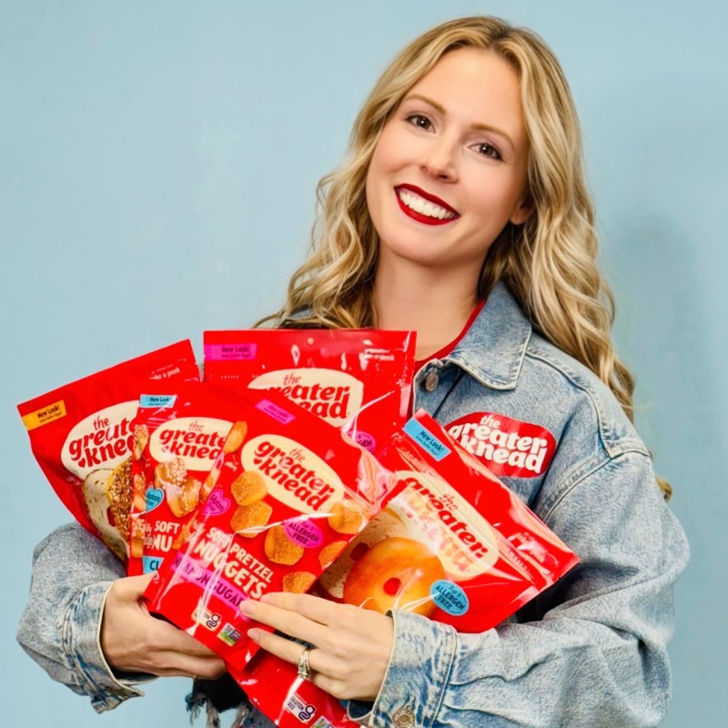 Advisor Michelle Carfango, CEO of gluten-free and allergy-friendly bakery The Greater Knead. She is smiling and holiding four bags of Great Knead Pretzel Nuggets against a light blue background.