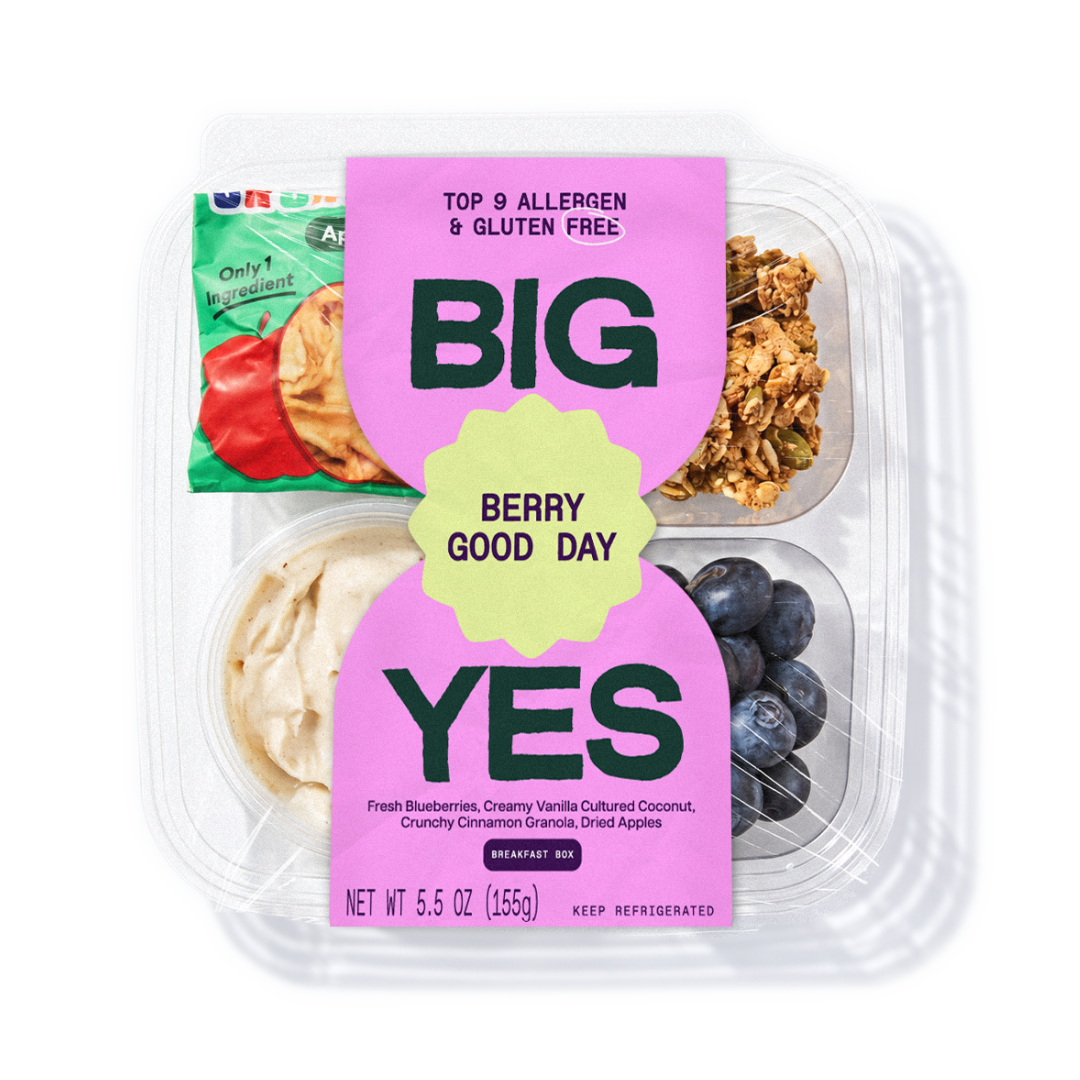 A clear plastic container of a breakfast box labeled 'BIG YES' with 'Berry Good Day' on a pink and yellow label. The box contains dried apples, granola, and blueberries, with a small packaged snack tucked behind the main container.