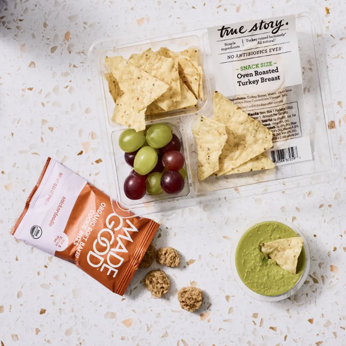 A snack box set from true story. & made good, featuring an oven roasted turkey breast snack size container with tortilla chips, green and red grapes, a bag of organic apple slices, and a small cup of green dip on a light speckled countertop.