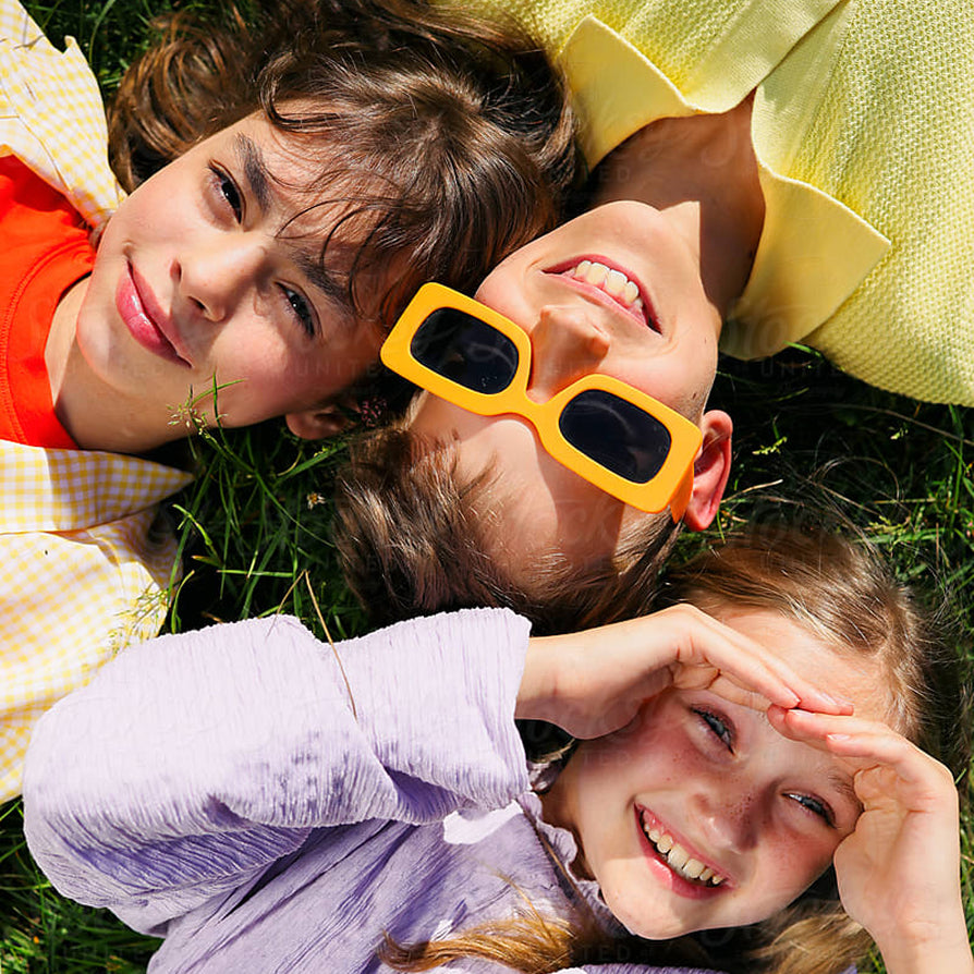 Three children lying on green grass outdoors, wearing bright yellow square sunglasses and casual colorful clothing.