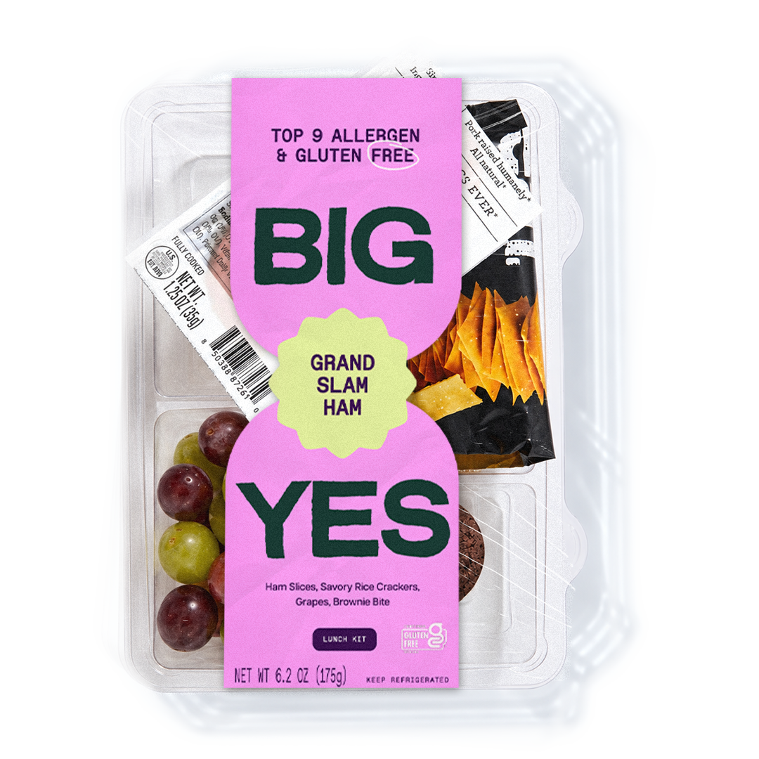 A clear plastic lunch kit container with a bright pink YES! brand label reading 'BIG GRAND SLAM HAM' and 'TOP 9 ALLERGEN & GLUTEN FREE.' Visible contents include ham slices, rice crackers, grapes, and banana bites arranged in compartments.