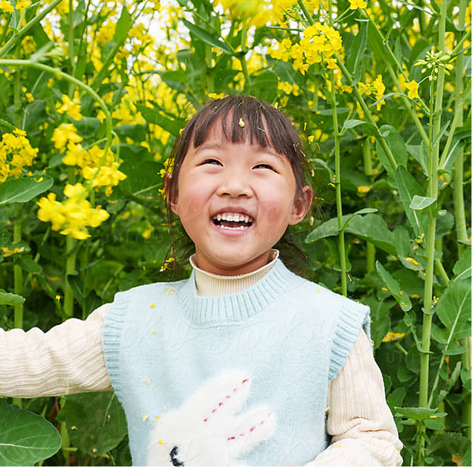A young girl with dark hair smiling joyfully in a field of yellow flowers. She is wearing a light blue knit sweater vest over a cream-colored long-sleeve shirt.