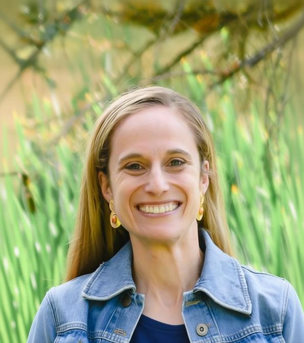A smiling woman with blonde hair wearing a light blue denim jacket and yellow earrings, photographed outdoors with blurred green plants and tree branches in the background.