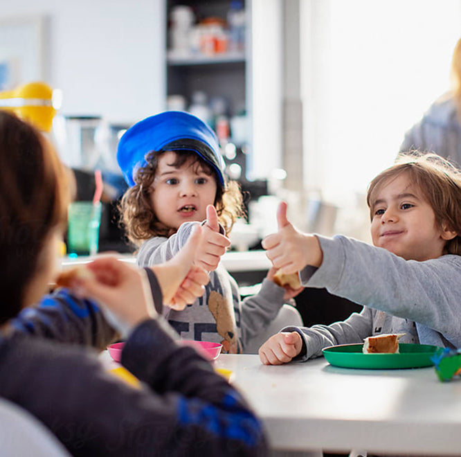 Two young children sitting at a table in a bright indoor setting, both giving a thumbs-up gesture about their lunch food.